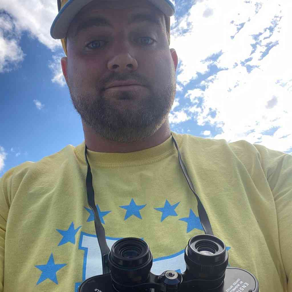Jesse Heubel, a technology teacher and wrestling coach, in a selfie, wearing a yellow shirt and binoculars around his neck.